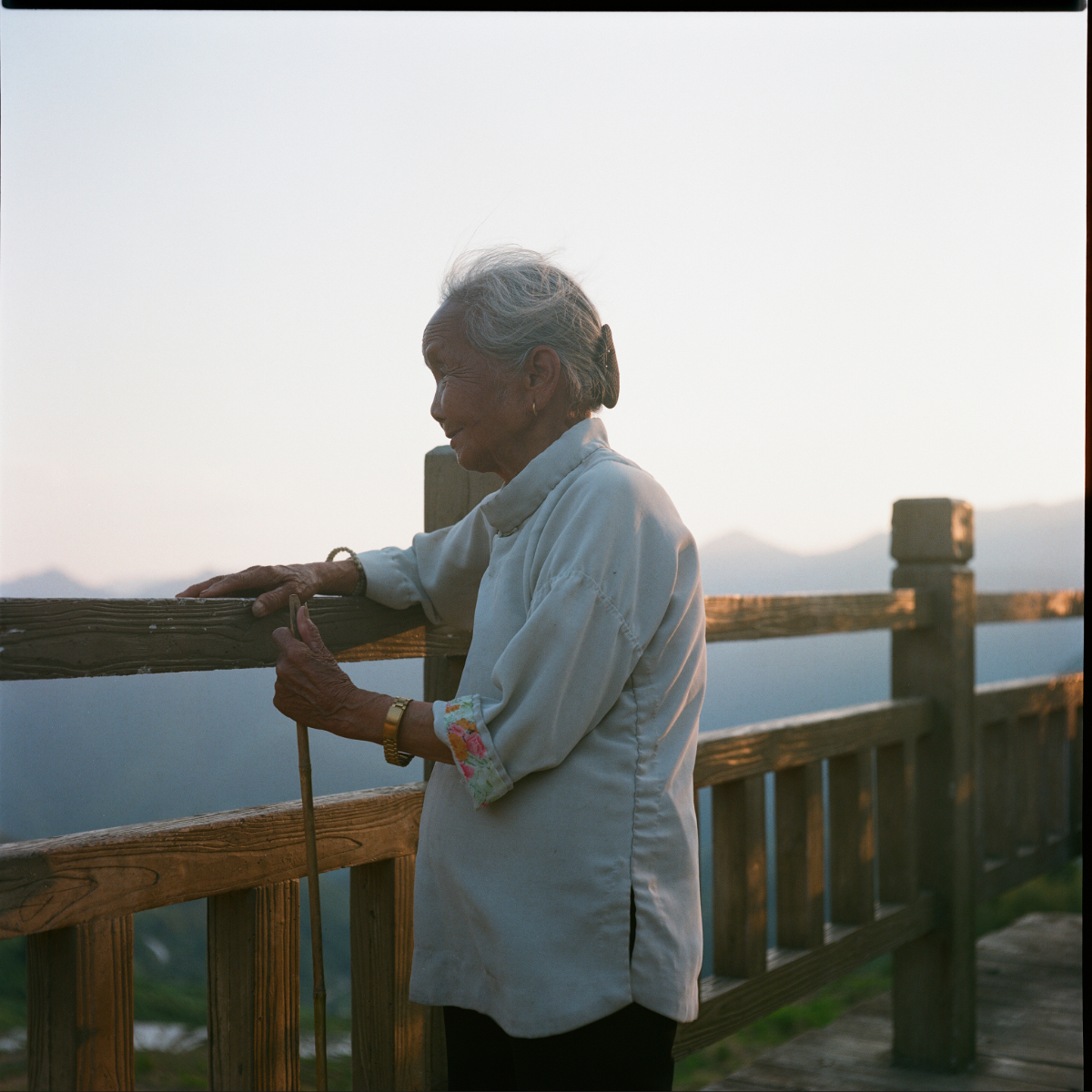 An elderly woman leaning on a wooden railing at sunset